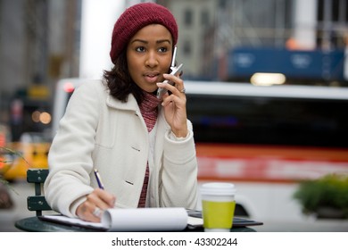 A mobile business woman in the city talks on her cell phone while writing something down in her notepad.