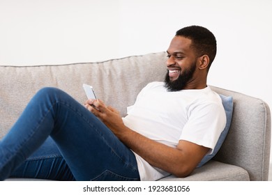 Mobile Communication. Black Guy Using Smartphone Browsing Internet And Texting Lying On Couch At Home, Side View. Mobile Phone User Networking Using Social Media Application.