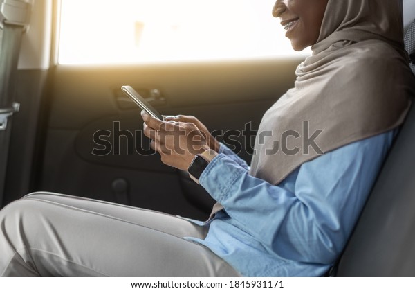 Mobile\
Communication. Black Muslim Businesswoman In Hijab Using Smartphone\
On Backseat Of Car, Enjoying Road Drive With Private Driver, Going\
To Work With Taxi, Cropped Image, Side\
View