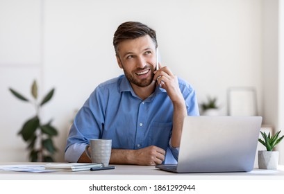 Mobile Communication. Handsome Male Entrepreneur Talking On Cellphone On Workplace In Office, Millenial Business Man Sitting At Desk With Laptop, Enjoying Pleasant Phone Conversation, Copy Space