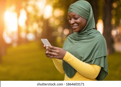 Mobile Communication. Portrait Of Smiling Black Muslim Lady In Hijab Using Smartphone Outdoors, Positive African Islamic Woman Texting With Friends Or Browsing Internet While Standing On Autumn Street