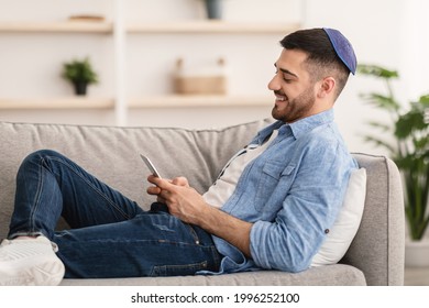 Mobile Communication. Side view of happy young jewish man using modern smartphone device while sitting on sofa at home, modern design interior. Cheerful guy typing an sms message at social network