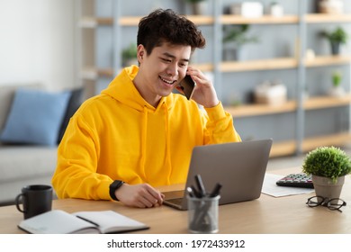 Mobile Communication. Smiling Asian Male Freelancer Talking On Cellphone And Working On Laptop At Home Office, Positive Man Using Computer And Having Pleasant Phone Conversation, Enjoying Distance Job