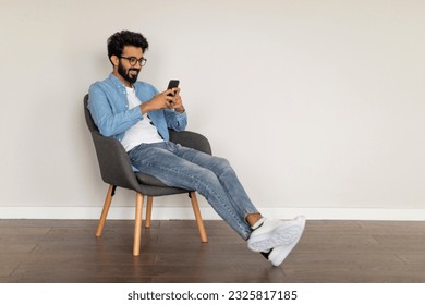 Mobile Communication. Young Indian Guy Messaging On Smartphone While Relaxing In Armchair At Home, Happy Handsome Eastern Male Texting With Friends Or Browsing Social Media, Copy Space - Powered by Shutterstock