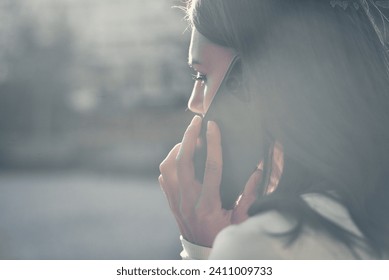 Mobile Conversation. Side view close up of a young beautiful woman which engaged in a phone call, holding a modern smartphone to her ear in a naturally lit outdoor environment.