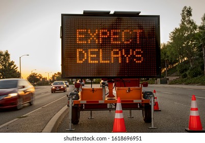 Mobile Electronic Traffic Sign stating “expect Delays” taken at sunset with traffic blurred driving past the sign and traffic cones - Shutterstock ID 1618696951