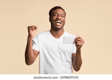 Mobile Games. Happy African American Man Holding Cellphone Shaking Fists Standing In Studio Over Yellow Background. - Powered by Shutterstock