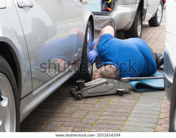 A mobile\
mechanic lies on a driveway looking underneath the chassis and\
bodywork of a silver sports car.A trolley jack is visible.His\
reflection is on the side of the\
vehicle