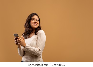 Mobile offer. Arab lady holding smartphone and looking back at free space, standing over brown background with empty space. Young woman checking new app promo