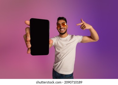 Mobile Offer. Cool Young Arab Guy In Sunglasses Pointing At Cellphone With Empty Black Screen, Recommending New App Or Website, Offering Space For Your Ad In Neon Light, Mockup. Selective Focus