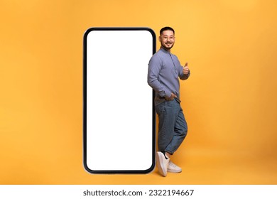 Mobile Offer. Excited asian guy leaning on big smartphone with blank screen and showing thumb up gesture, millennial man recommending great new app or website, yellow background, mockup - Shutterstock ID 2322194667