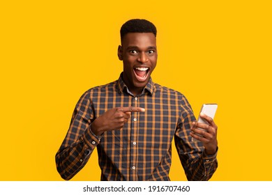 Mobile Offer. Excited Black Guy Pointing At Smartphone In His Hands, Emotionally Reacting To New App, Overjoyed Millennial African American Man Standing Isolated Over Yellow Background, Copy Space