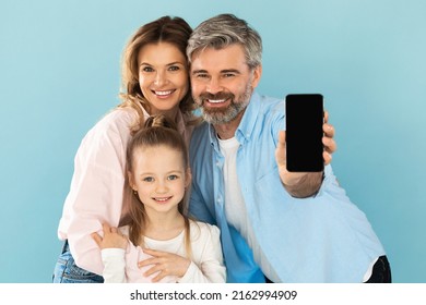 Mobile Offer. Family Showing Smartphone With Blank Screen Posing Embracing Smiling To Camera Standing On Blue Background In Studio. Great Application Advertisement. Mockup