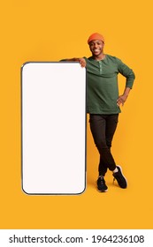 Mobile Offer. Happy Black Guy Leaning At Big Smartphone With Blank White Screen, Cheerful African Man Demonstrating Copy Space For Your Design Or Advertisement, Posing Over Yellow Background, Mockup