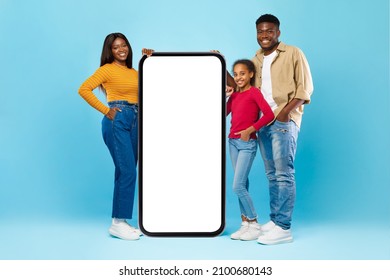 Mobile Offer. Happy Casual Black Family Leaning On Big Huge Cell Phone With Empty White Screen, Recommending Cool New App Or Website Offering Free Copy Space For Your Ad, Mock Up, Blue Studio Wall