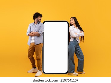 Mobile offer. Millennial indian couple leaning on giant smartphone with mockup, advertising or promoting app or website over yellow studio background