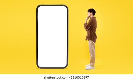 Mobile Offer. Thoughtful Guy Looking At Big Smartphone With Blank Screen, Thinking About Innovative Application Standing On Yellow Background. Man Posing Near Huge Gadget. Studio Shot, Panorama - Shutterstock ID 2310010897