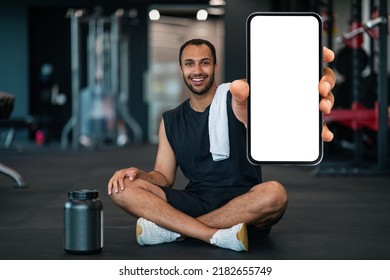 Mobile Offer. Young Black Sportsman Demonstrating Big Blank Smartphone At Camera, Smiling African American Male Athlete Sitting On Floor At Gym And Showing Cellphone With White Screen, Mockup