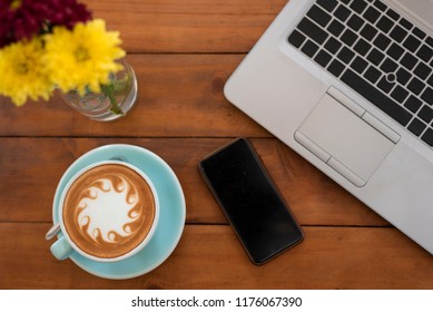 A Mobile Phone And Blue Cup Of Coffee Have A Abstract Shape Milk Froth Put On Wooden Table And Have Brew Spoon Put Near A Cup,have A Blurry Yellow Flower And Laptop Computer In Top View Coffee Concept