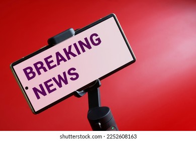Mobile phone with breaking news inscription on gimbal selfie stick. Concept of breaking and sensational news, live broadcasting, video blogging, streaming and journalism. Red background. Close-up - Shutterstock ID 2252608163