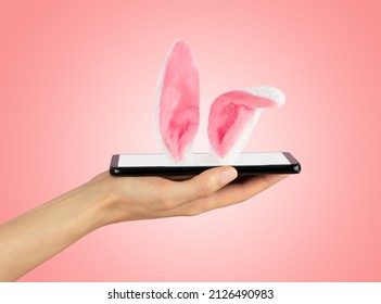 Mobile phone is lying on female palm, Easter Bunny ears sticking out of smartphone screen. Soft pink background. Happy Easter Concept. - Shutterstock ID 2126490983