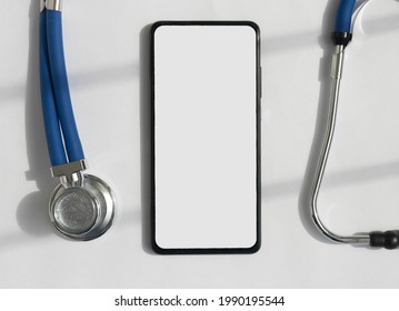 Mobile Phone Mockup With Stethoscope Over White Desk, Top View. Template With White Screen Of Smartphone For Medical App.