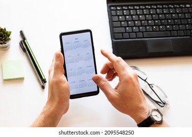 Mobile Phone With An Open Calendar For 2022 Year In A Male Hand On A Working Desk. In The Hand Of A Mobile Phone With A Calendar For 2022 Year. On The Desktop A Laptop, Two Pens, Glasses And Note