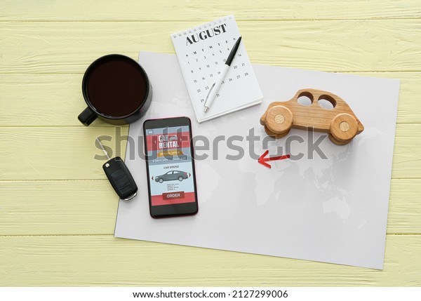 Mobile phone with open car rent\
app, cup of coffee, calendar and toy on yellow wooden\
background