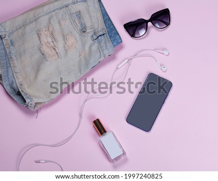 Mobile phone, sunglasses, white headphones, hipster denim shorts for a girl, perfume in a glass bottle on a light pink background. View from above