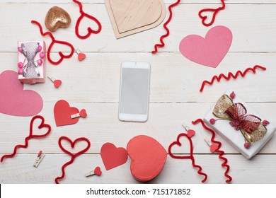 Mobile phone surrounded by cards, decorations and presents, top view. Love and holiday concept. Valentines day symbols: paper, wooden and fuzzy wire hearts, pegs on white vintage background. - Powered by Shutterstock