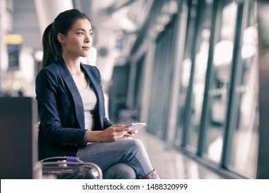 Mobile Phone Travel Businesswoman Waiting In Airport Lounge With Hand Luggage. Young Asian Woman On Business Trip Using Cell Smartphone. Tech Device For Wifi Entertainment Online In Terminal.