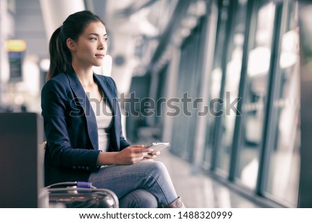 Mobile phone travel businesswoman waiting in airport lounge with hand luggage. Young Asian woman on business trip using cell smartphone. Tech device for wifi entertainment online in terminal.