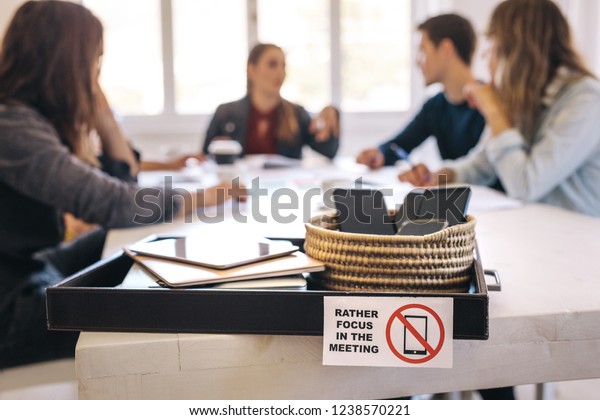 Mobile phones, laptops and digital tablets in a
tray on meeting table with business people sitting at back and
discussing work. Meeting without technology. A method for more
productive meetings