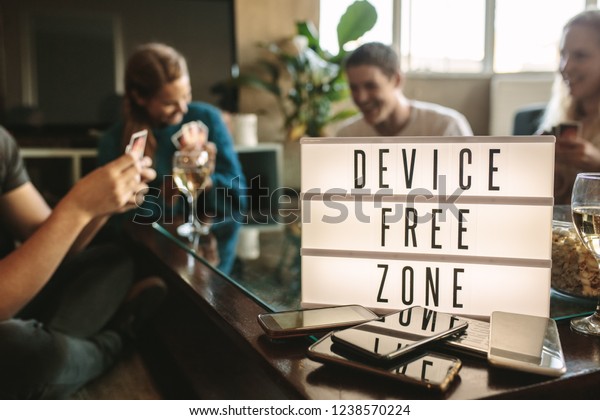 Mobile\
phones on table with device free zone sign. Group of young men and\
woman hanging out together at no phone\
zone.