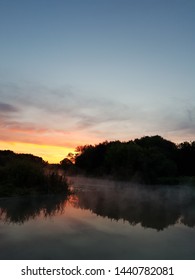 Mobile Photography. Sunrise Over The River. Haze Of Mist Over The Water. Reflection Of Dark Trees, Shades Of Crimson Sky In River Water. The Sun Glow In The Background In The Lower Third Of The Image.