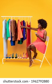 Mobile Shopping. African American Female Using Smartphone Choosing Clothes Sitting Near Clothing Rack Posing Over Yellow Background. Shopping App Concept. Studio Shot, Vertical