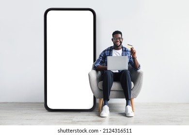 Mobile Shopping App. Black Man Sitting Near Big Phone With Empty Screen, Using Laptop And Credit Card Over Grey Wall, Mockup. Device With Space For App Or Website Design