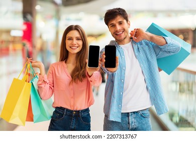 Mobile Shopping App. Happy Buyers Couple Showing Cellphones Advertising App Smiling To Camera Holding Shopper Bags Posing In Modern Mall Indoor. Ecommerce And Technology Concept. Mockup