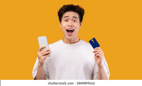 Mobile Shopping App. Portrait Of Joyful Asian Guy Holding And Showing Credit Card And Cellphone Posing Standing Over Orange Studio Background. Excited Male Teen Making Online Purchase