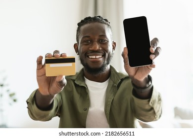 Mobile Shopping Appliation. Happy Black Guy Holding Credit Card In Hand And Showing Smartphone With Blank Screen To Camera, Free Space For Mock Up, Digital Pay