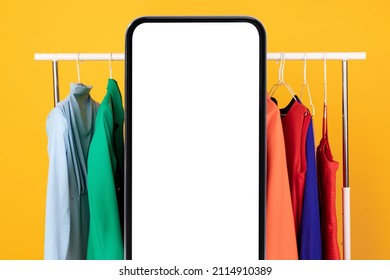 Mobile Shopping Application. Big Smartphone With Blank Screen Standing Near Clothing Rail With Colorful Jackets Over Yellow Studio Background. Fashion And Technology, E-Commerce App. Mockup