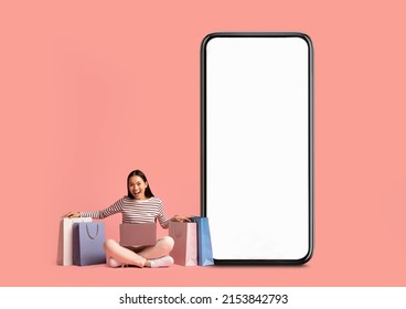 Mobile Shopping Application. Excited Asian Lady Showing Blank Empty Cell Phone Screen Sitting On Floor With Shopper Bags Isolated On Pink Studio Background. Online Order, Virtual Store Concept. Mockup