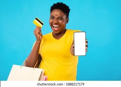 Mobile Shopping Application. Excited Black Woman Showing Smartphone Empty Screen Holding Credit Card Standing Over Blue Studio Background. Female Shopper Recommending App And Phone Banking. Mockup