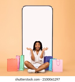 Mobile Shopping Application. Excited Black Lady Showing Blank Empty Cell Phone Screen Sitting On Floor With Shopper Bags Isolated Over Studio Background. Online Order, Virtual Store Concept. Mockup