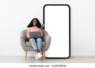 Mobile Shopping Application. Happy Black Lady Sitting On Chair Near Big Cellphone With Empty Screen Using Credit Card And Laptop, White Studio Wall Background, Mockup, Collage