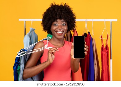 Mobile Shopping Application. Joyful African Female Showing Smartphone Empty Screen To Camera Recomending Fashion And Style Assistant App Posing Near Clothing Rack Over Yellow Studio Background. Mockup