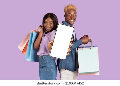 Mobile Shopping Application, Online Store. Happy Black Couple Showing Blank Empty Cell Phone Screen In Hand Holding Shopper Bags Standing Isolated On Purple Violet Studio Background. Mock Up Template