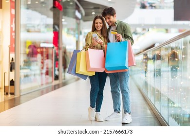 Mobile Shopping. Couple Of Buyers Using Phone And Credit Card Purchasing New Clothes In Application Holding Colorful Shopper Bags Standing In Modern Mall. Ecommerce Offers And Sales