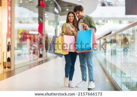 Mobile Shopping. Couple Of Buyers Using Phone And Credit Card Purchasing New Clothes In Application Holding Colorful Shopper Bags Standing In Modern Mall. Ecommerce Offers And Sales