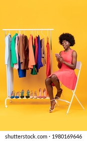 Mobile Shopping. Positive African American Lady Using Cellphone Purchasing Clothes Sitting Posing In Studio With Clothing Rack Over Yellow Background. Studio Shot, Vertical